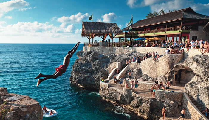 VibraGem Tours Rick's Cafe Day Tour in Jamaica with a man diving off a cliff into the blue ocean water.